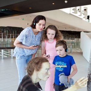 Een jong gezin (moeder met dochter en zoon) volgt samen een audiotour en bekijkt het kunstwerk van twee Romeinse vrouwen in Museum Hoge Woerd aandachtig.