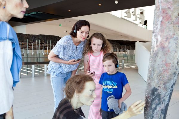 Een jong gezin (moeder met dochter en zoon) volgt samen een audiotour en bekijkt het kunstwerk van twee Romeinse vrouwen in Museum Hoge Woerd aandachtig.