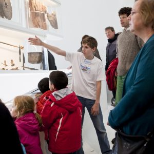 Een jonge rondleider vertelt met armgebaren iets over de museumstukken in de glazen vitrines en een groep bezoekers van diverse leeftijden kijkt en luistert aandachtig.