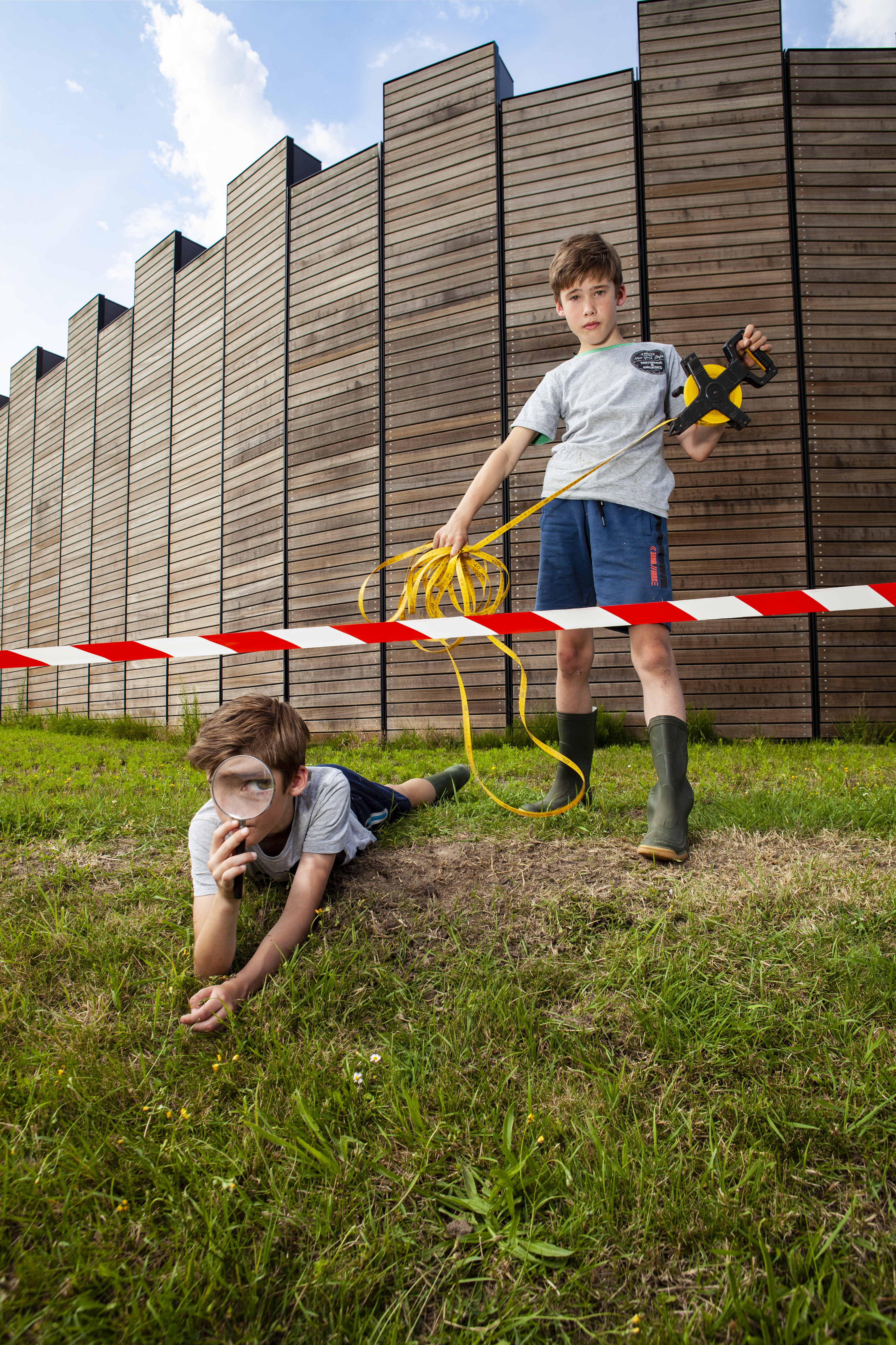 Castellum-promo-kinderen-vergrootglas-meetlint-afzetlint Een jongen ligt op de grond voor de muren van het castellum en houdt een vergrootglas voor zijn gezicht, de andere jongen houdt een meetlint vast.
