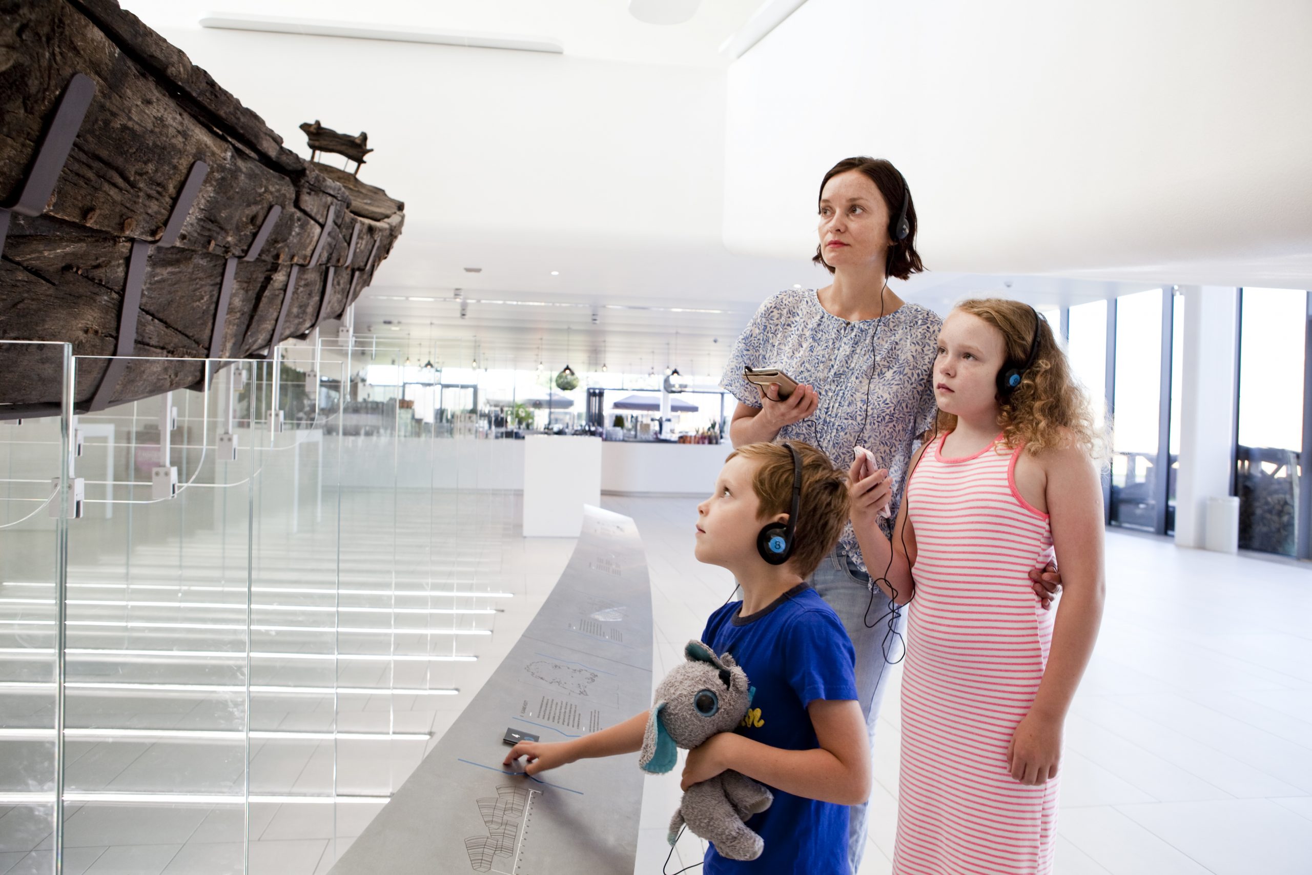 Museum-hoge-woerd-audiotour-gezin-romeins-schip-(c)AafkeHolwerda Een jong gezin (moeder met dochter en zoon) volgt samen een audiotour en bekijkt het opgegraven Romeinse schip aandachtig.