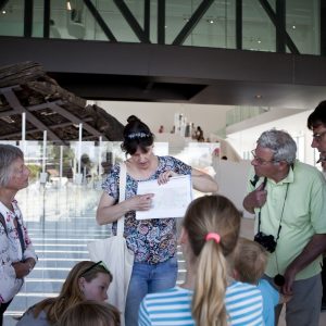 Een vrouwelijke gids wijst informatie aan op een hand-out. Een groep geïnteresseerde bezoekers van verschillende leeftijden kijkt en luistert aandachtig naar haar.