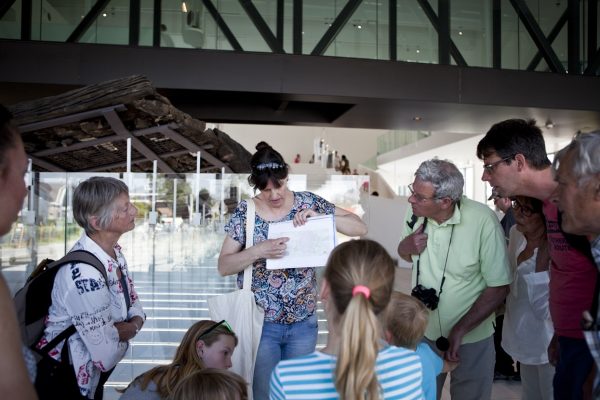 Een vrouwelijke gids wijst informatie aan op een hand-out. Een groep geïnteresseerde bezoekers van verschillende leeftijden kijkt en luistert aandachtig naar haar.