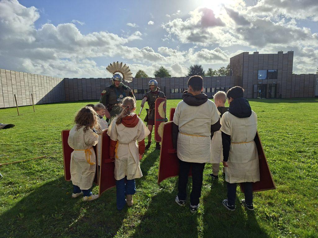 Heb jij het in je om een Romeinse soldaat te worden? Langs de Limes, de noordgrens van het Romeinse Rijk, stonden 2000 jaar geleden forten vol legionairs die dag en nacht trainden. Tijdens deze actieve workshop ontdek je hoe dat eraan toeging én mag je het zelf proberen.
Je leert de basis van het zwaardvechten en verdedigen: eerst op een oefenpaal, daarna met een mede‑soldaat en uiteindelijk in twee groepen. Natuurlijk oefen je ook met de werpspeer, een onmisbaar wapen voor elke legionair. Kun jij in formatie werpen zonder je eigen troepen te raken? Tot slot train je als echte eenheid: marcheren, draaien, reageren, want in het Romeinse leger stond de veiligheid van de groep altijd voorop.
Ontmoet echte Romeinse soldaten
Bij de wachttoren staan onze grensbewakers op de uitkijk. Ze dragen replica‑uitrusting, vertellen over het leven van een legionair en beantwoorden graag al je vragen. Een unieke kans om de Romeinse Limes‑soldaten van dichtbij te ontmoeten.